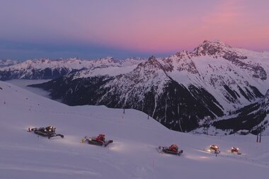 Mehrere Pistenraupen der Gargellner Bergbahnen präparieren bei Sonnenuntergang die schneebedeckten Hänge des Skigebiets. Der Himmel leuchtet in zarten Rosa- und Lilatönen, während sich die umliegenden Berge majestätisch im Hintergrund erheben. Eine friedliche Winterstimmung in der hochalpinen Landschaft von Gargellen. | © Montafon Tourismus GmbH, Marie Schilcher