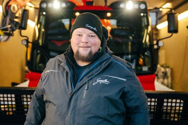 Ein Mitarbeiter der Gargellner Bergbahnen steht lächelnd in der Werkstatt vor einer roten Pistenraupe. Er trägt eine dunkelblaue Jacke und eine schwarze Mütze mit dem Schriftzug „Gargellen“. Im Hintergrund ist die beleuchtete Fahrzeughalle mit der Pistenraupe zu sehen. | © Montafon Tourismus GmbH, Marie Schilcher