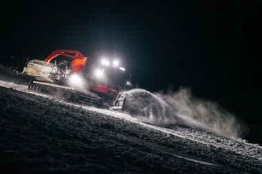 Eine Pistenraupe der Gargellner Bergbahnen ist nachts im Einsatz und schiebt frischen Schnee auf die Piste. Im Schein der hellen Scheinwerfer wirbeln Schneekristalle auf und glitzern in der Dunkelheit – ein beeindruckender Moment technischer Präzision und winterlicher Stimmung in Gargellen. | © Montafon Tourismus GmbH, Marie Schilcher