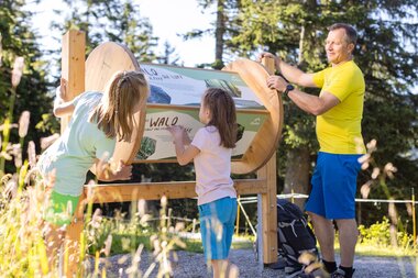 Ein Vater mit zwei Kindern auf Golmis Forschungspfad. Sie drehen gerade an einer rechteckigen Informationstafel mit vielen Informationen zum Wald. | © Golm Silvretta Lünersee Tourismus GmbH Bregenz, Stefan Kothner