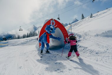 Maskottchen Golmi vom Erlebnisberg Golm mit einem Kind, dass am skifahren ist | © Golm Silvretta Lünersee Tourismus GmbH Bregenz, Philipp Schilcher